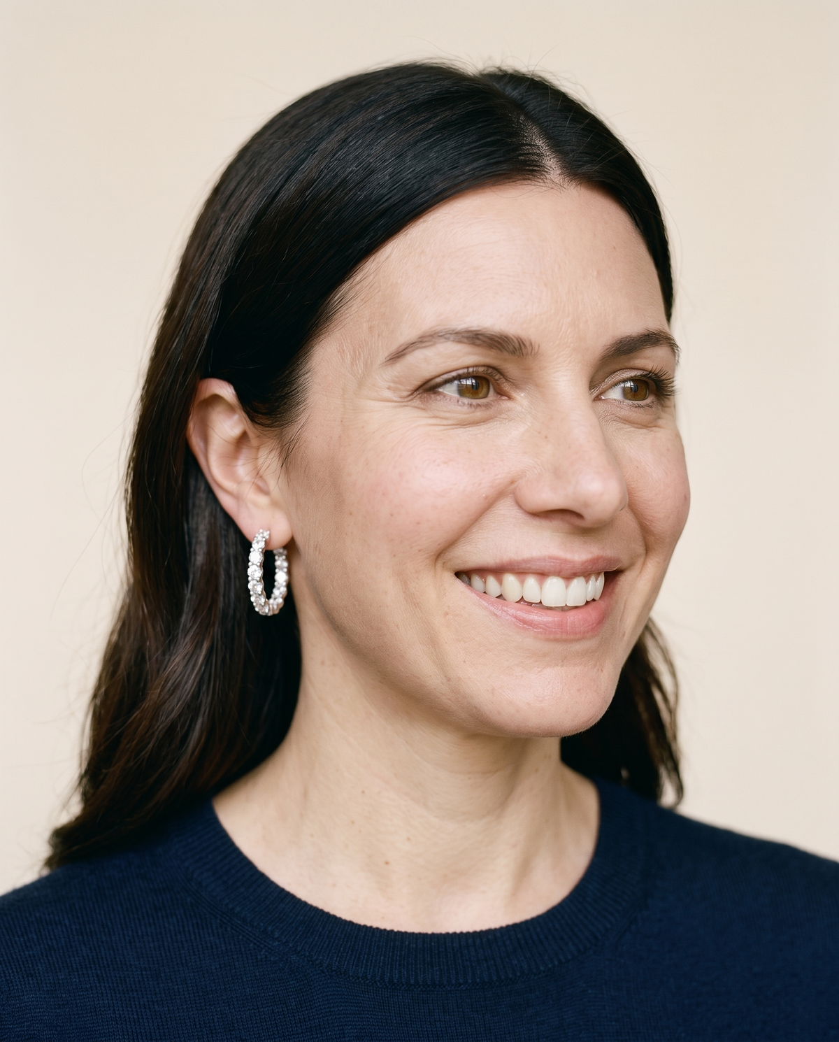 Smiling woman with dark hair wearing moissanite pavé hoop earrings and a navy knit